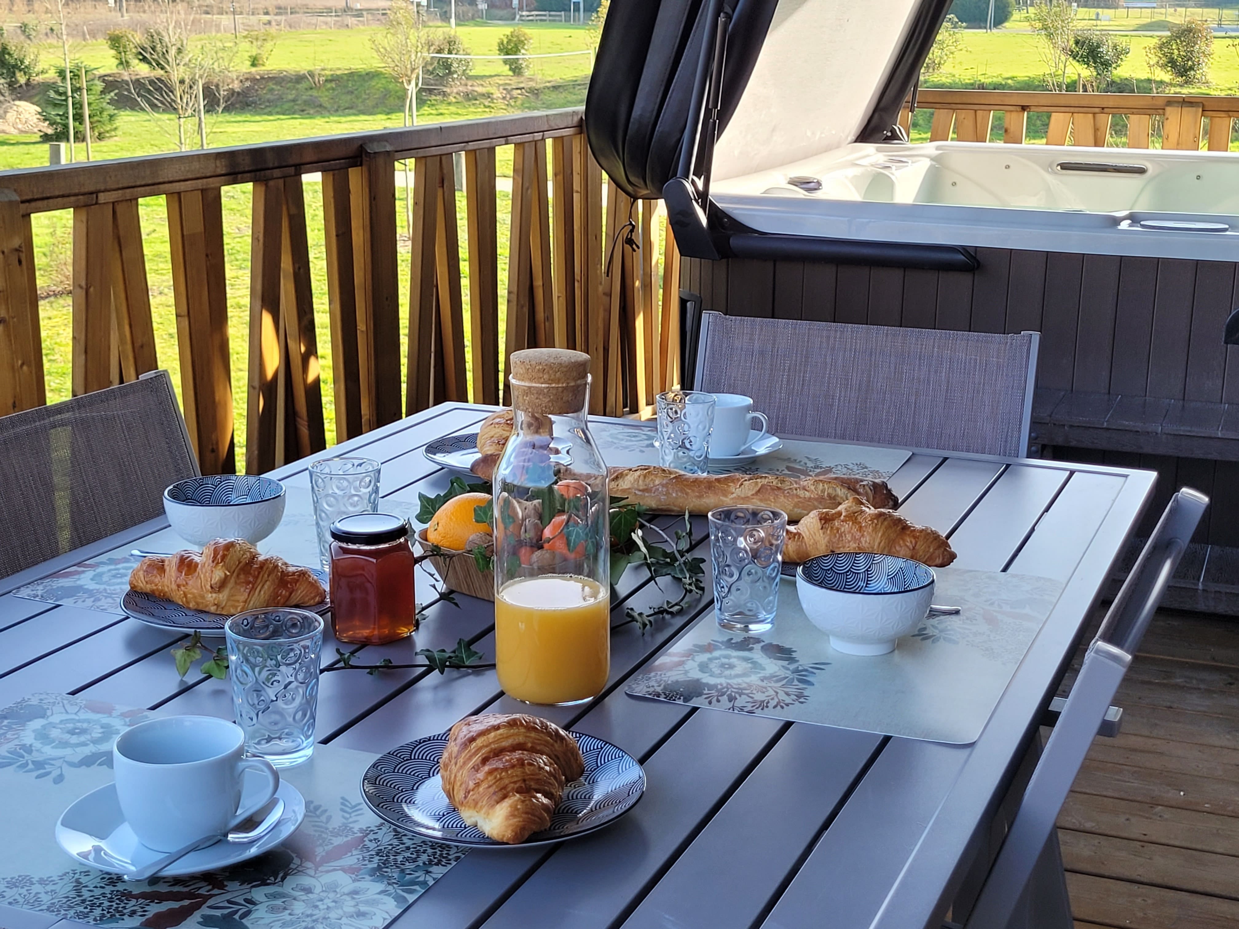 Terrasse ensoleillée avec vue sur un paysage verdoyant et spa en arrière-plan, une table dressée pour un petit-déjeuner gourmand avec des produits frais.